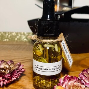 May include: A small glass bottle filled with a yellow liquid and herbs. The bottle has a black dropper top and a white label that reads "Prosperity in the bag!"