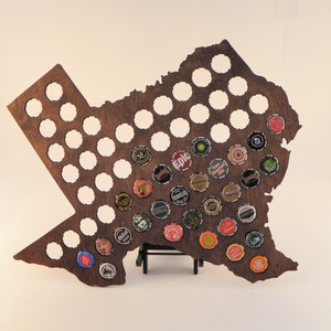 May include: A wooden cutout of the state of Texas with a stand, designed to hold bottle caps. The cutout is filled with various bottle caps, some with logos and some with designs.