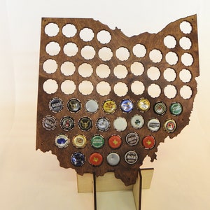 May include: A wooden Ohio-shaped bottle cap holder with numerous circular openings. Several bottle caps are inserted, showcasing various colorful designs. The holder is supported by a wooden base, creating a decorative display for beer bottle caps.
