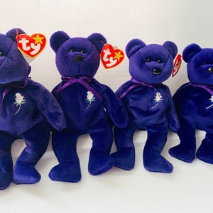 May include: Four plush purple teddy bears with black button eyes, a white rose, and a purple ribbon around their necks. Each bear has a Ty tag attached. The bears are sitting on a white surface.