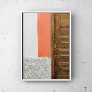 May include: A framed photograph of a wooden door with a peach-colored wall behind it. The door is made of wood panels and has a simple metal handle. The wall is painted in a light peach color and has a white stripe running down the side.