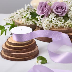 May include: A roll of lavender ribbon with a white floral pattern is placed on a stack of wooden slices. The ribbon is unspooled, showing its satin texture. The background features purple roses and white flowers, suggesting a craft or gift-wrapping theme.