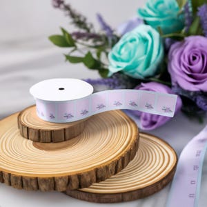 May include: A roll of pink and white patterned ribbon with floral designs. The ribbon is on a wooden spool and is sitting on a set of wooden slices.