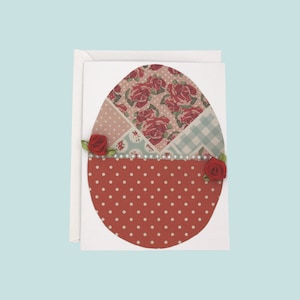 以下が含まれることがあります： 手作りのイースターカード。卵の形をしており、上半分は花柄や模様のパッチワーク、下半分は白い水玉模様の赤色です。小さな赤いバラとイミテーションパールで飾られています。