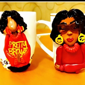 Puede incluir: Dos tazas de cerámica con figuras decorativas. Una figura lleva un suéter rojo con la inscripción "PRETTY BROWN" en amarillo. La otra figura lleva gafas de sol y pendientes amarillos con el símbolo de la paz. Ambas figuras tienen pelo oscuro y pintalabios rojo.