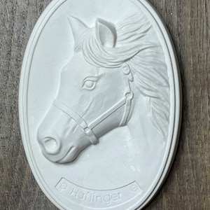 May include: White oval ceramic plaque featuring a raised relief sculpture of a horse's head. The horse is wearing a bridle and has a flowing mane. The plaque is inscribed with the word "Haflinger".