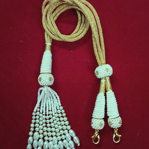 Könnte beinhalten: Eine weiße Perlenkette mit goldenen Akzenten und Quasten. Die Kette besteht aus kleinen weißen Perlen und hat eine goldene Schnur. Die Quasten bestehen aus weißen Perlen und haben goldene Akzente.