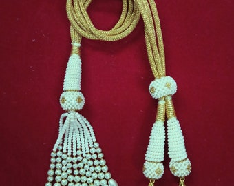 Peut inclure: Un collier de perles blanches avec des accents dorés et des glands. Le collier est fait de petites perles blanches et a un cordon doré. Les glands sont faits de perles blanches et ont des accents dorés.