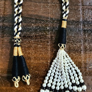 May include: Two black and white braided cords with gold accents and tassels. One tassel is made of white pearls and the other is made of black thread.