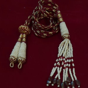 May include: A long, maroon and gold cord with white pearl tassels and brown beads. The cord has gold accents and a red stone on each tassel.