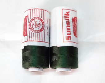 May include: Two spools of dark green embroidery thread. The spool on the left has a white label with red text that reads "A Product By Komal Textiles, India, Art No. 1202". The spool on the right has a white label with red text that reads "Sunsilk, Computer Embroidery Thread".