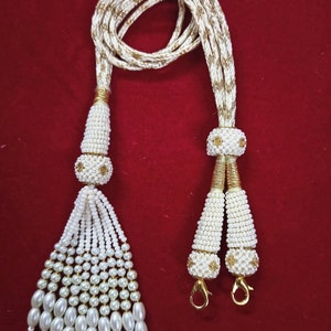 Może przedstawiać: Biały naszyjnik z koralików z złotymi akcentami i dwoma frędzlami. Naszyjnik wykonany jest z małych białych koralików i ma złoty łańcuszek ze złotym zapięciem.