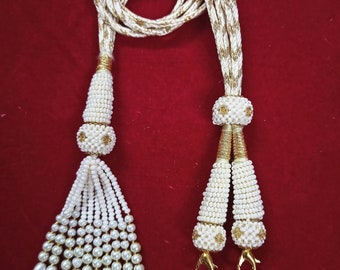Peut inclure: Un collier de perles blanches avec des accents dorés et deux glands. Le collier est fait de petites perles blanches et a une chaîne dorée avec un fermoir doré.