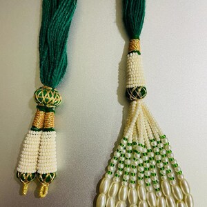 May include: Two green and white beaded tassels with gold accents. The tassels are made of small white beads and green beads. The tassels are attached to green cords.