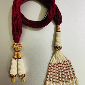 Puede incluir: Un collar de cuentas marrones y blancas con detalles dorados. El collar tiene dos borlas con cuentas blancas y detalles rojos.