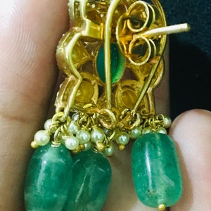 K&ouml;nnte beinhalten: Goldfarbene Ohrringe mit smaragdgr&uuml;nen, tropfenf&ouml;rmigen Steinen und kleinen wei&szlig;en Perlen. Die Ohrringe haben ein florales Design mit einem Steckerverschluss. Die Ohrringe werden in einer Hand gehalten.