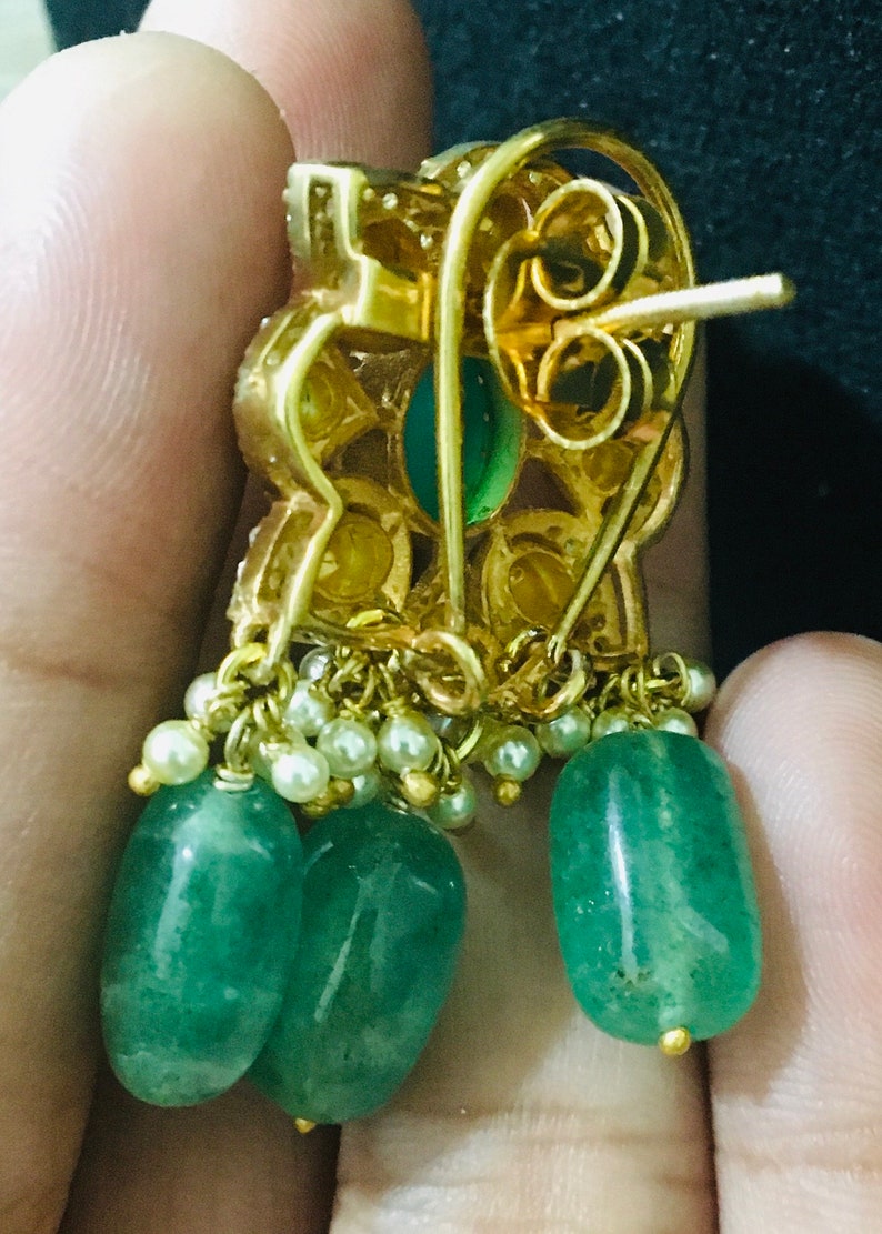 K&ouml;nnte beinhalten: Goldfarbene Ohrringe mit smaragdgr&uuml;nen, tropfenf&ouml;rmigen Steinen und kleinen wei&szlig;en Perlen. Die Ohrringe haben ein florales Design mit einem Steckerverschluss. Die Ohrringe werden in einer Hand gehalten.