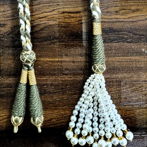 May include: Two green and gold tassel cords with white accents. One tassel is made of white pearls and the other is a simple tassel.