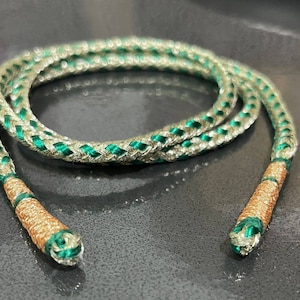 Puede incluir: Un cordón trenzado con un diseño verde y plateado. El cordón tiene extremos marrones.