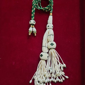 以下が含まれることがあります： 緑と金のアクセントが付いた白いビーズのタッセルネックレス。ネックレスには、緑と金のコードが付いたタッセルが2つ付いています。