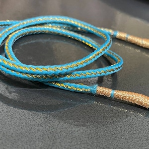 Puede incluir: Un cordón azul y dorado con un diseño trenzado. El cordón está doblado por la mitad y los extremos están atados.