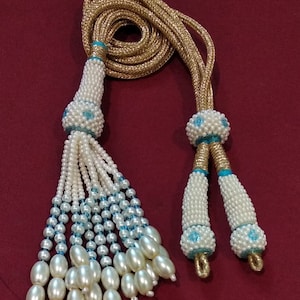 May include: A gold cord necklace with two white pearl tassels. Each tassel has a blue and white bead at the top and a cluster of white pearl beads at the bottom.