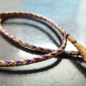 May include: A braided cord necklace with gold and purple thread. The cord is twisted and knotted, creating a unique design.