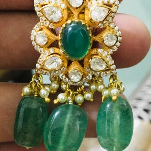 K&ouml;nnte beinhalten: Goldfarbene Ohrringe mit floralem Design, mit einem zentralen ovalen gr&uuml;nen Edelstein und tropfenf&ouml;rmigen klaren Steinen. Drei gro&szlig;e, ovale gr&uuml;ne Perlen h&auml;ngen darunter, akzentuiert mit kleinen wei&szlig;en Perlen.