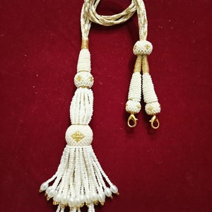 Puede incluir: Collar de borlas de cuentas blancas con detalles dorados. El collar tiene un cordón largo y delgado con dos borlas en los extremos. Las borlas están hechas de cuentas blancas y tienen una cuenta dorada en la parte superior.