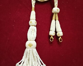 Peut inclure: Collier à glands en perles blanches avec des accents dorés. Le collier a un cordon long et fin avec deux glands aux extrémités. Les glands sont faits de perles blanches et ont une perle dorée au sommet.