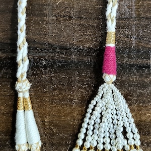 May include: Two white braided cords with gold accents and different tassel ends. One tassel is made of white beads and the other is a pink and white tassel.