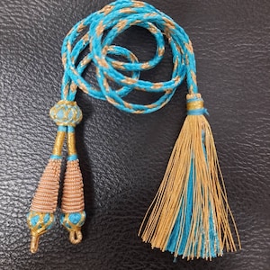 Może przedstawiać: Sznur z frędzlami w kolorze niebieskim i złotym, z dwoma frędzlami na jednym końcu i jedną frędzlą na drugim końcu.