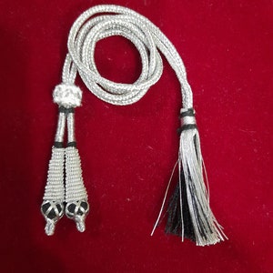 May include: A silver cord with two tassel ends. One end has a silver bead and the other end has a black and silver tassel.
