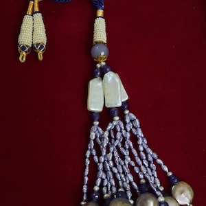 Op de afbeelding: Een blauwe en witte kralenkwast met een parel- en paarse steendesign. De kwast hangt aan een blauw koord en heeft een witte parelzoom.