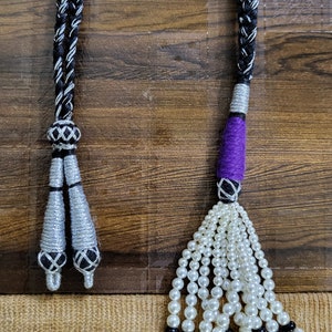 May include: Two black and silver braided cords with tassels. One tassel is silver and the other is purple with a white pearl fringe.
