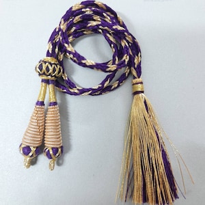 Puede incluir: Cordón trenzado morado y dorado con dos extremos de borla y una bola decorativa en la parte superior.