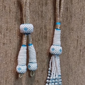 May include: Two white beaded tassels with blue accents. The tassels are attached to gold cords. The tassels are on a brown textured background.