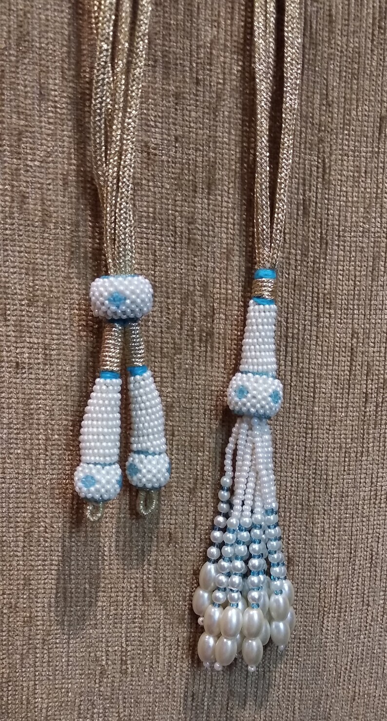 May include: Two white beaded tassels with blue accents. The tassels are attached to gold cords. The tassels are on a brown textured background.