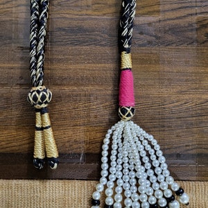 May include: Two black and gold braided cords with tassels. One tassel is made of white pearls and black beads, the other is a pink and gold tassel.