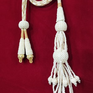 Peut inclure: Un pompon blanc en perles avec un cordon en fil doré. Le pompon a plusieurs brins de perles qui se terminent par de petits bouquets de perles.