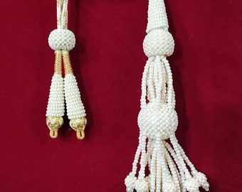 Peut inclure: Un pompon blanc en perles avec un cordon en fil doré. Le pompon a plusieurs brins de perles qui se terminent par de petits bouquets de perles.