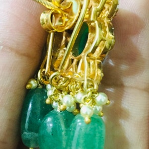 K&ouml;nnte beinhalten: Goldfarbene Ohrringe mit einem h&auml;ngenden Design. Jeder Ohrring hat eine Ansammlung kleiner wei&szlig;er Perlen und drei gr&uuml;ne, tropfenf&ouml;rmige Edelsteine. Die Ohrringe haben eine dekorative Fassung im Filigranstil.