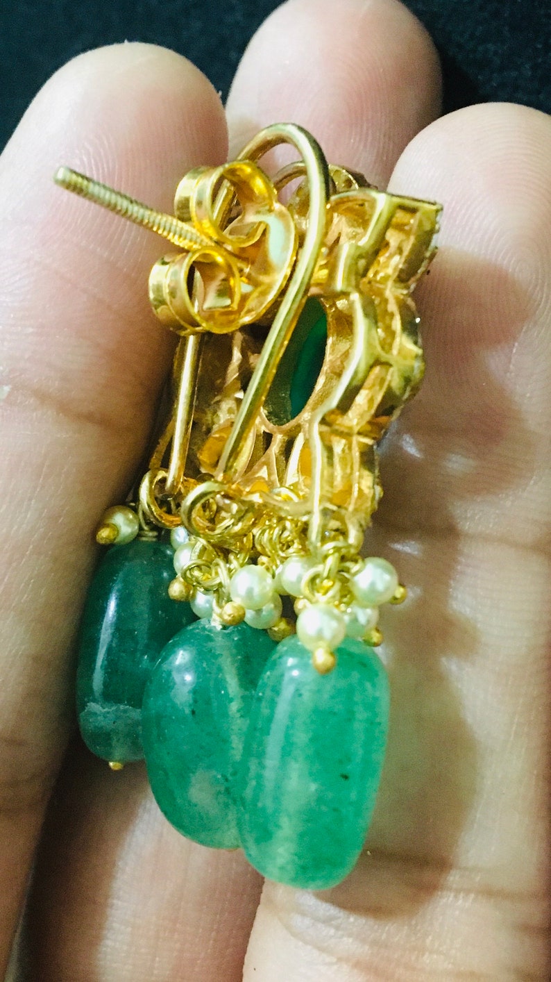 K&ouml;nnte beinhalten: Goldfarbene Ohrringe mit einem h&auml;ngenden Design. Jeder Ohrring hat eine Ansammlung kleiner wei&szlig;er Perlen und drei gr&uuml;ne, tropfenf&ouml;rmige Edelsteine. Die Ohrringe haben eine dekorative Fassung im Filigranstil.