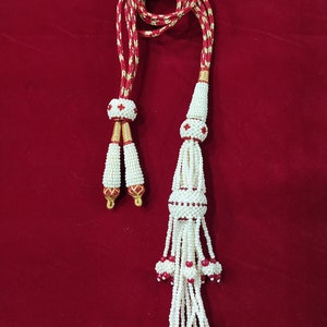 Peut inclure: Un collier de perles blanches avec des accents rouges et dorés. Le collier a un pompon à l'extrémité et est fait de petites perles rondes.