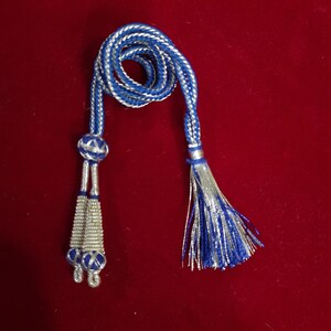 Könnte beinhalten: Eine blaue und silberne Schnur mit Quasten an jedem Ende. Die Schnur ist gedreht und hat einen silbernen Faden, der durch sie verläuft. Die Quasten bestehen aus blauen und silbernen Fäden.