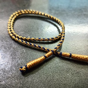 May include: A gold and blue braided cord with a knotted end. The cord is lying on a grey surface.