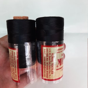 May include: Two spools of black thread wrapped in clear plastic. The label reads "Komal Textiles" with contact information for the company.