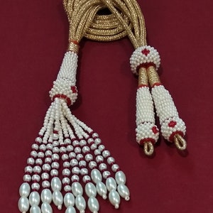 Puede incluir: Un collar de cuentas blancas y doradas con detalles rojos. El collar tiene dos borlas con cuentas blancas y detalles rojos.