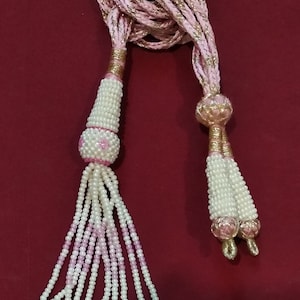 May include: A decorative pink and white beaded tassel with a braided cord. The tassel features white and pink beads, with a gold-coloured accent. The tassels are arranged in a cascading pattern, with a large oval bead at the end of each strand.
