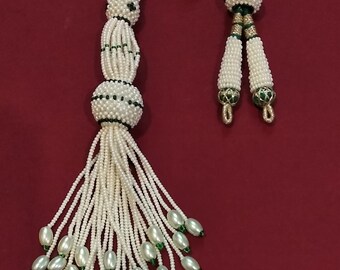 Peut inclure: Un collier de perles blanches avec des accents verts et un cordon vert et or. Le pompon est fait de petites perles blanches et a une grosse perle blanche au sommet. Le cordon est fait d'un fil vert et or.